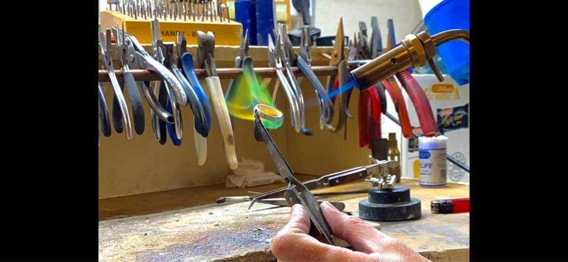 Aan de werkbank wordt een ring met een soldeervlam bewerkt, met tangen en gereedschap aan het rek erachter.